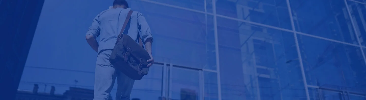 A man walking towards a glass building while carrying a bag.